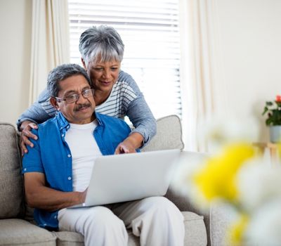 Senior man sitting on couch with spouse standing behind him both looking at laptop