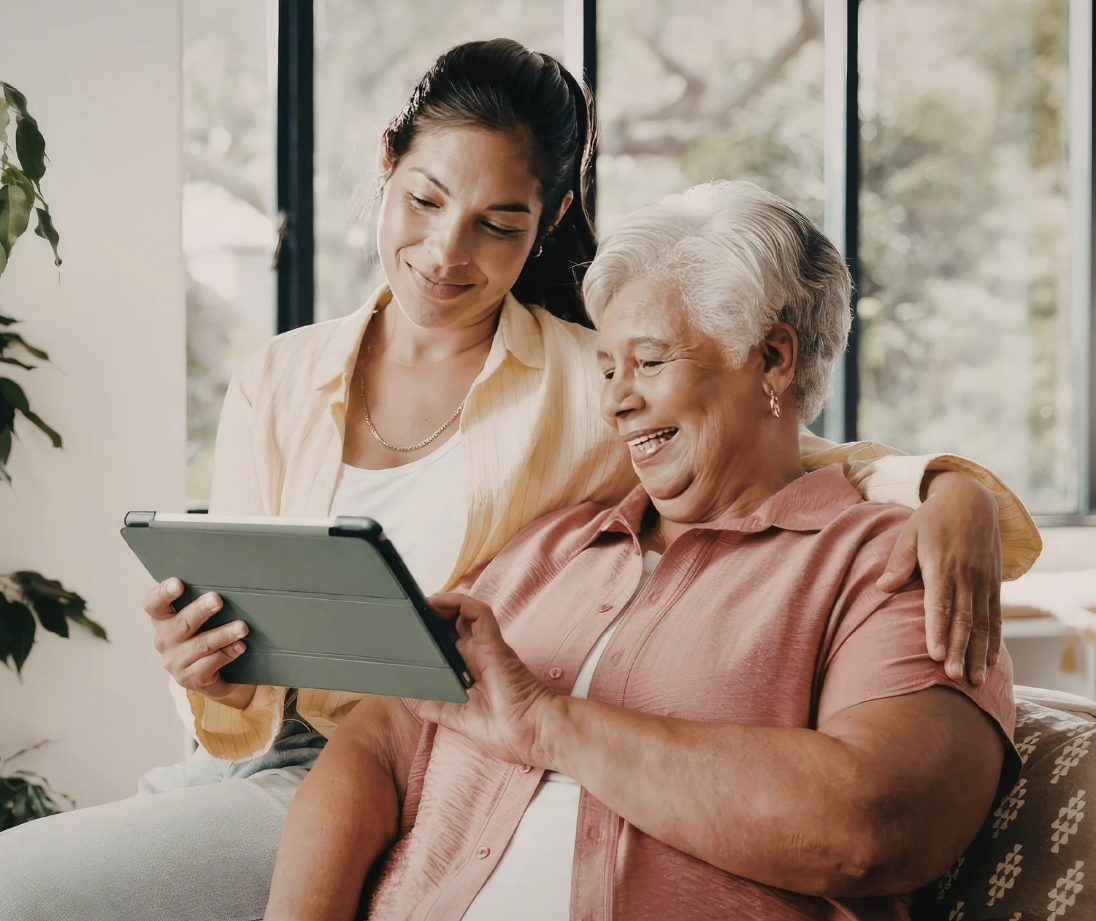 A younger woman and an older woman smile while looking at a tablet together on a couch, highlighting connection, technology, and intergenerational support in a bright home setting.