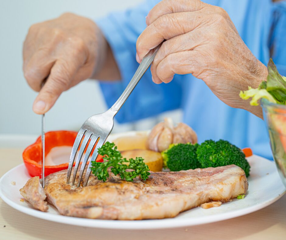 Older adult cutting grilled chicken served with broccoli, carrots and salad.