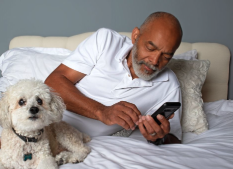 senior man laying across bed looking at cell phone white dog beside him