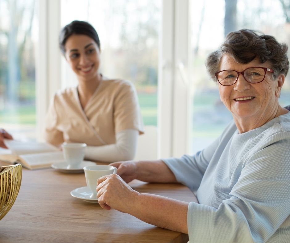 young woman having coffee and reading with senior woman in retirement community