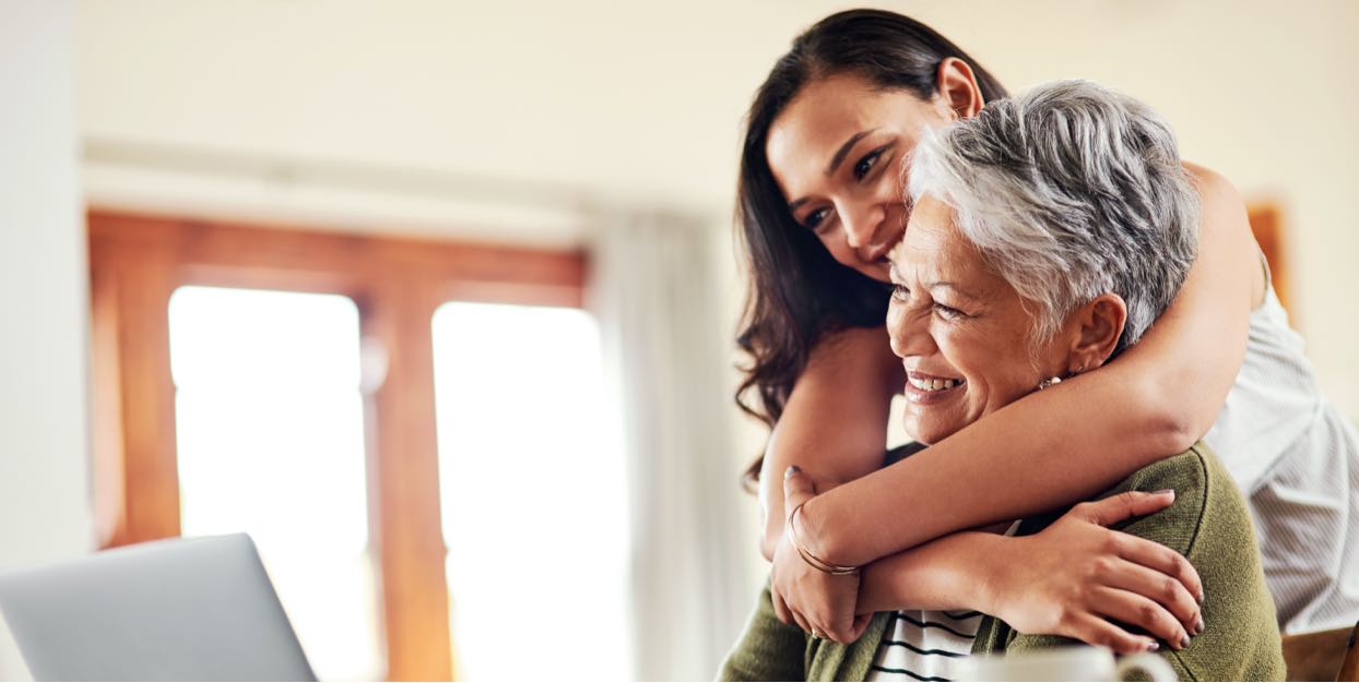 adult child hugging senior mom while both look at laptop