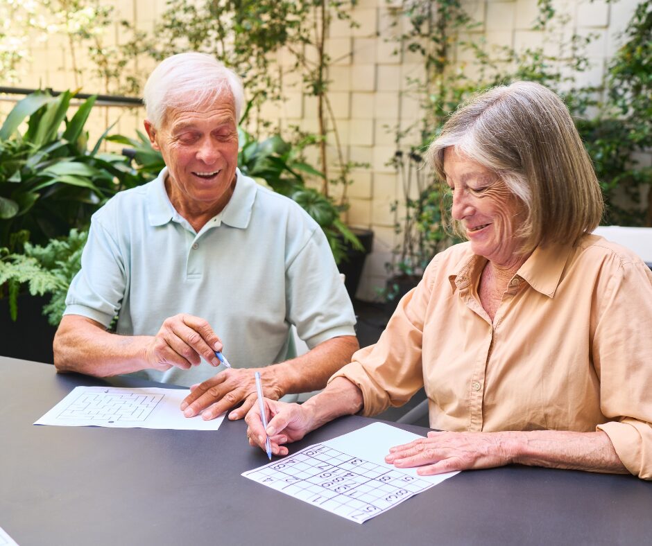 Older man and woman smiling while completing crossword puzzles together at an outdoor table.