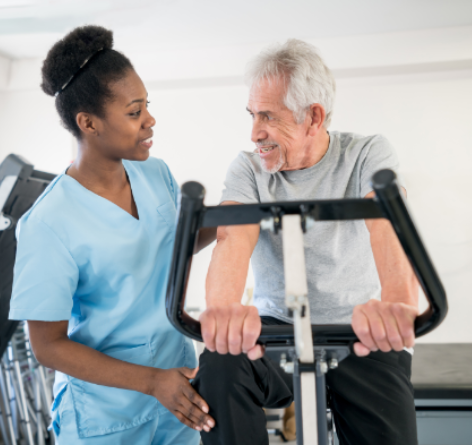 senior man using an exercise bike while looking at physical therapy assistant who is helping him recover