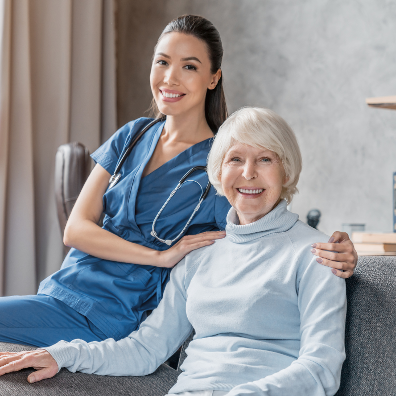 Senior woman in blue turtleneck sitting on couch with female nurse next to her